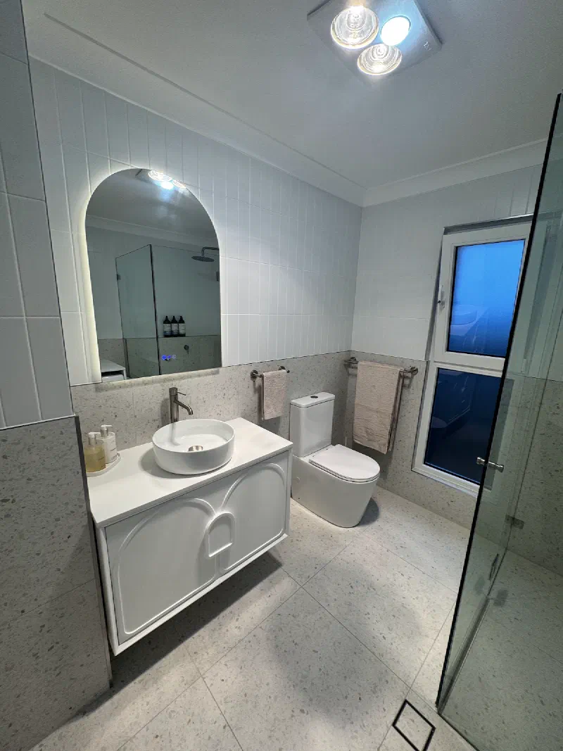 Bathroom with a white vanity and round basin, an arched mirror with a wall light, a toilet, towel rails and a glass shower screen.