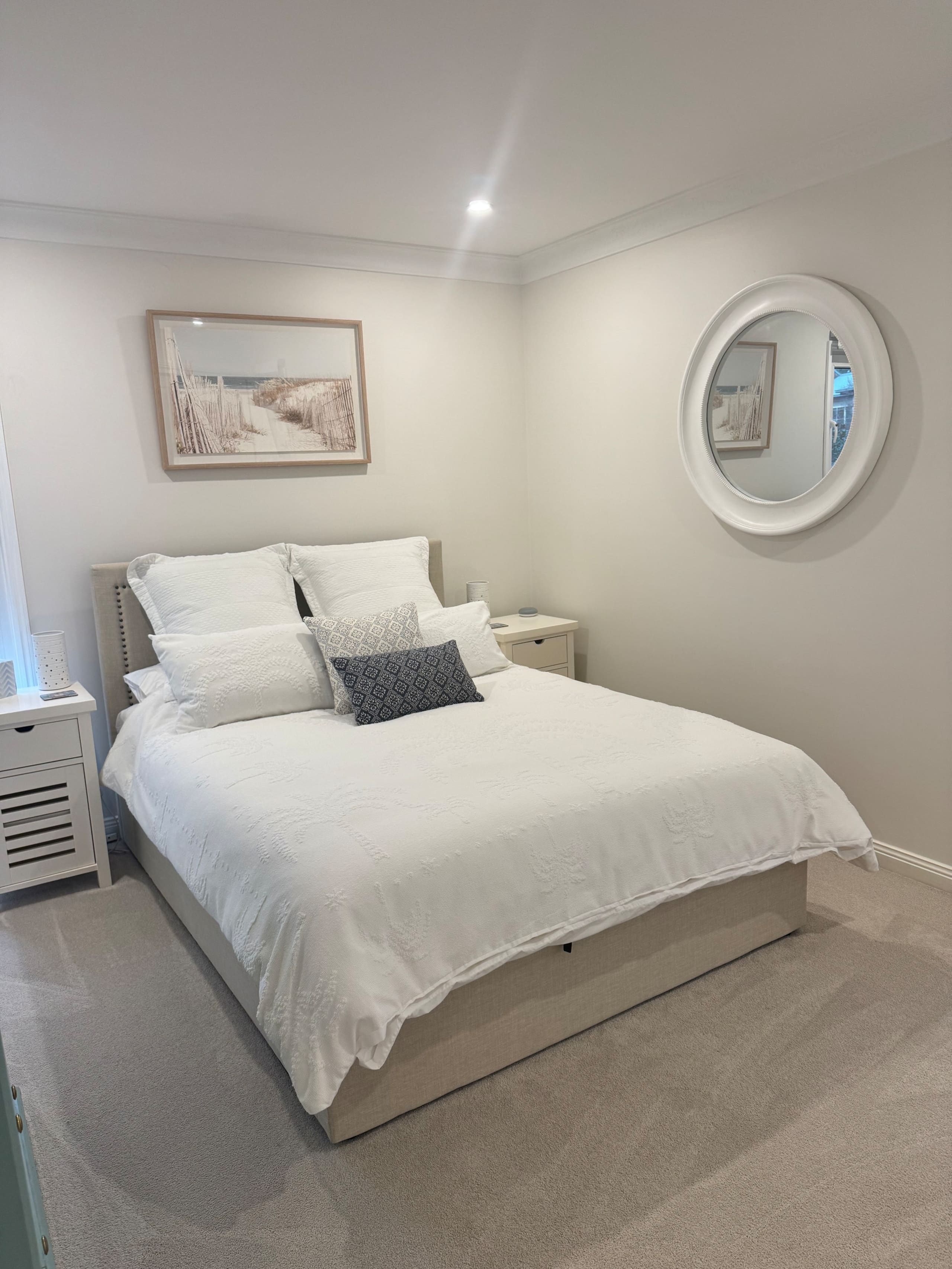 Queen bed with white linen and cushions, a framed coastal print, a round mirror and bedside tables