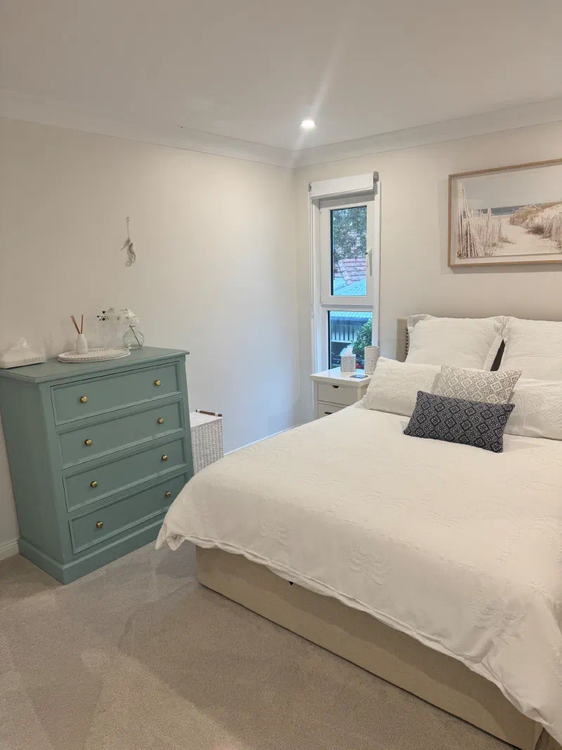 Queen bed with white linen beside a sage-green chest of drawers, with a window looking onto the garden.