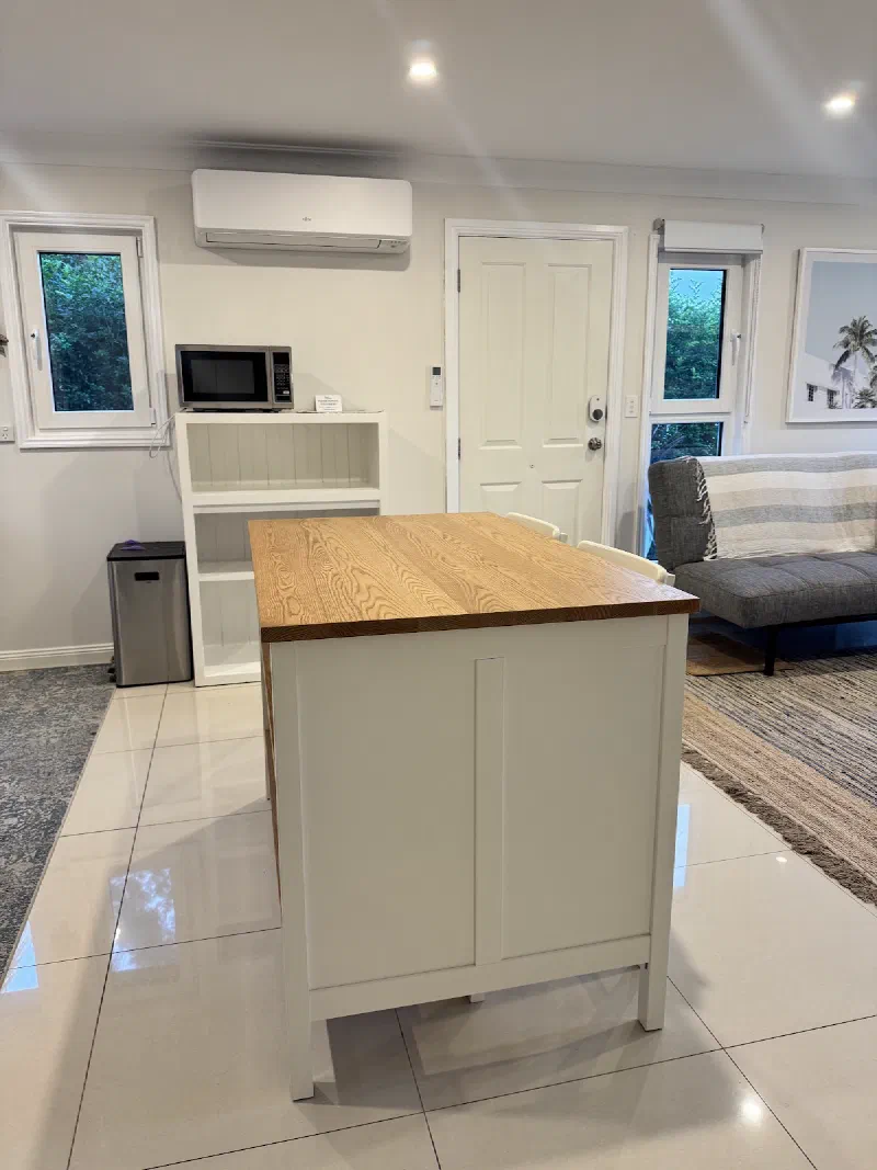 Living and dining area with a timber-topped island bench, a grey sofa, open shelving with a microwave and a wall-mounted air conditioner.