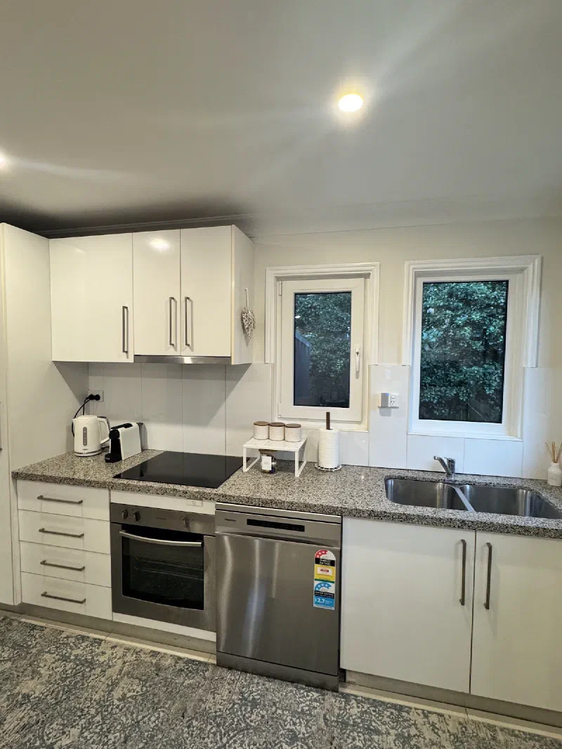 Kitchen wall with white cabinets, a black cooktop, stainless steel oven and dishwasher, and a sink beneath two windows.