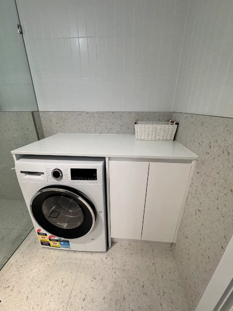 Front-loading washing machine beside a white cupboard in the tiled laundry, with a woven basket on the bench.