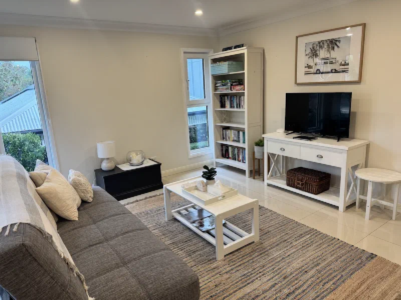 Living room with a grey sofa, white coffee table and striped rug, a television on a white console, a tall bookshelf and a framed coastal print.