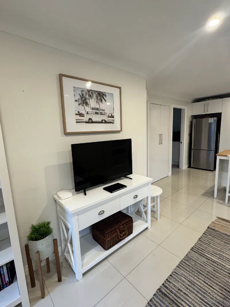 Television on a white console beneath a framed coastal print, with drawers below and a view through to the kitchen.